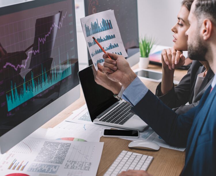 Side view of data analysts working with charts on paper and computer monitors at table